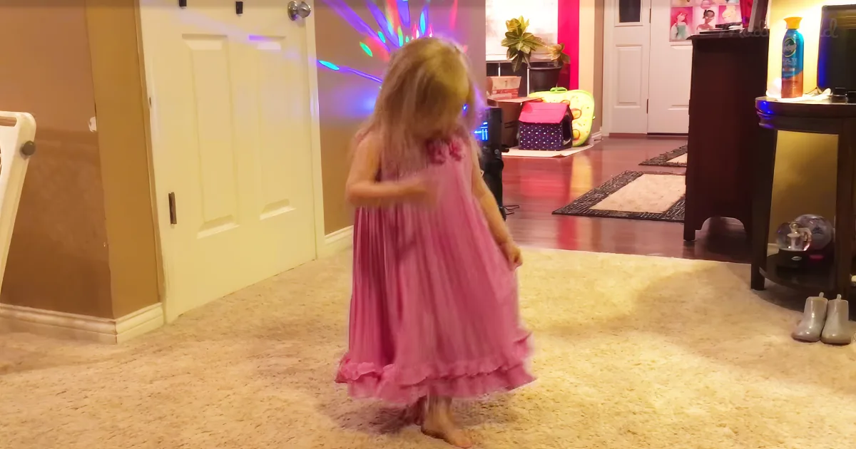 A young girl in a pink dress dances barefoot on a carpeted floor with colorful lights in the background.