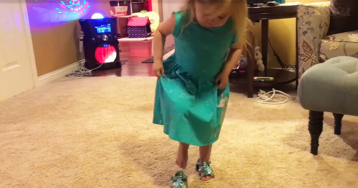 A young girl in a teal dress stands on a carpeted floor, holding the hem of her dress, with a colorful arcade machine and...