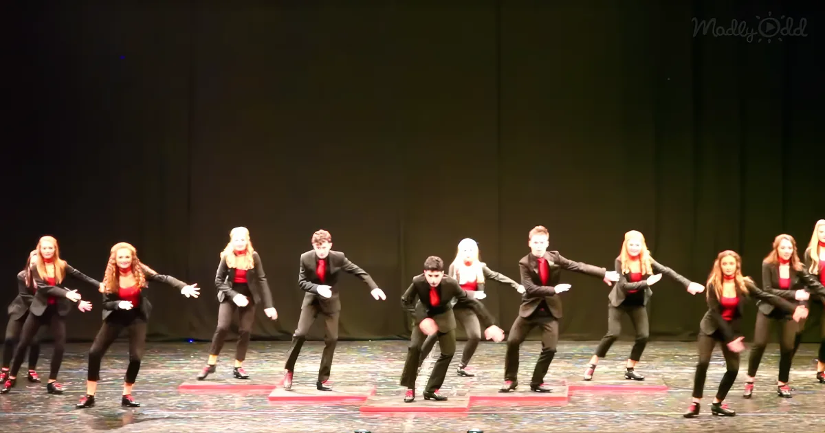 A group of dancers in black suits and red tops perform synchronized moves on stage.
