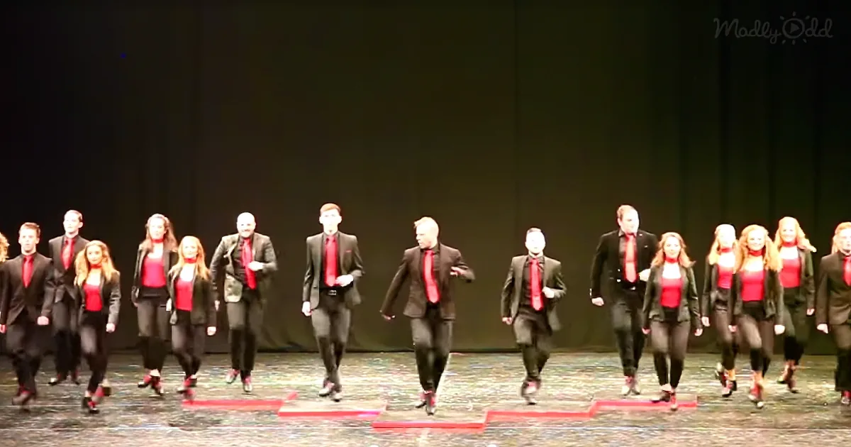 A group of performers in black suits and red shirts walk in unison across a stage.
