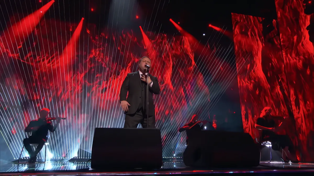 A man in a suit sings into a microphone on a stage with red lighting and dynamic visuals.