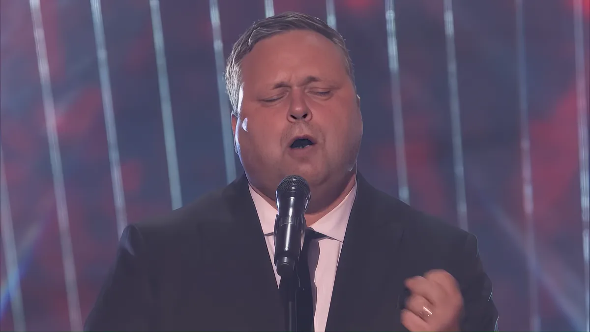A man in a suit sings passionately into a microphone on stage with a colorful background.