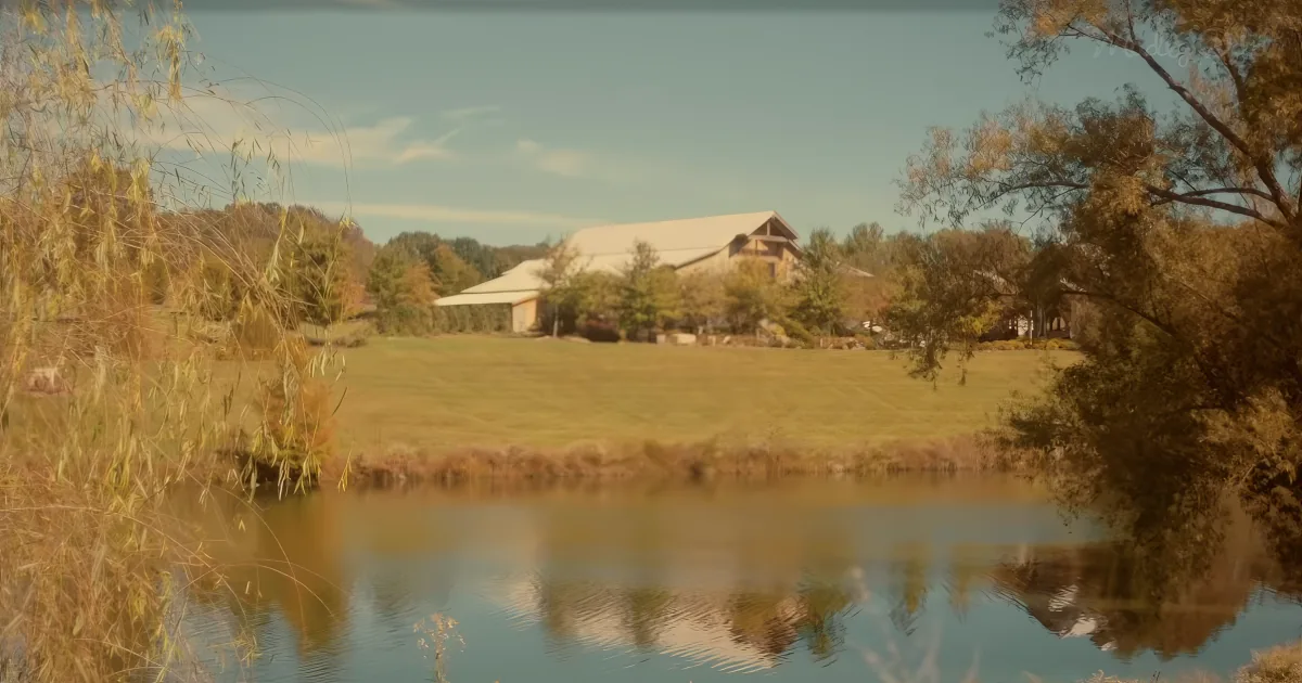 A serene pond reflects a barn and surrounding trees under a clear sky.