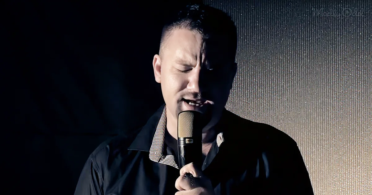 A man in a dark jacket sings into a microphone, his eyes closed, against a textured background.