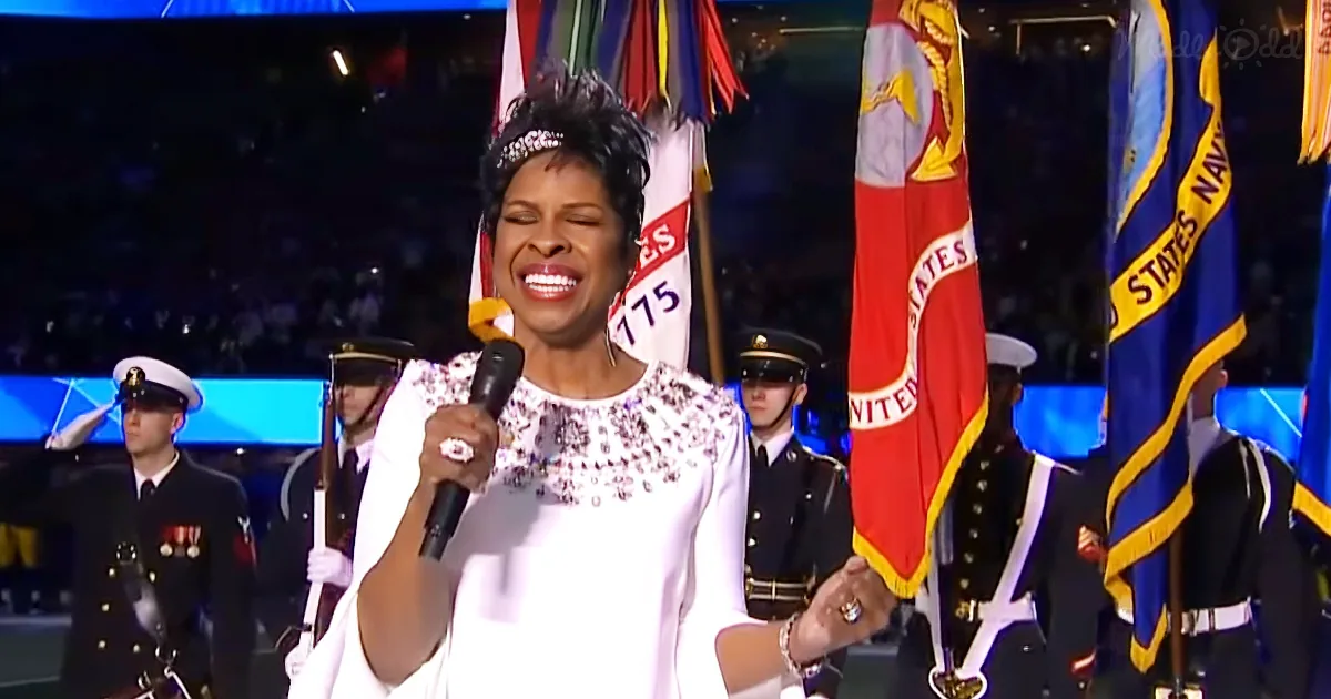 A woman in white attire sings into a microphone, surrounded by military personnel and flags.