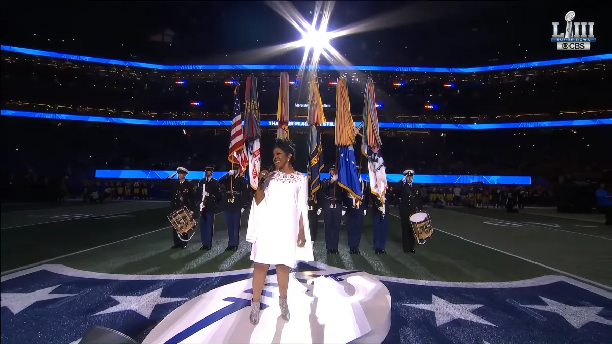 A woman in a white dress stands on a stage with flags and drummers in a stadium.