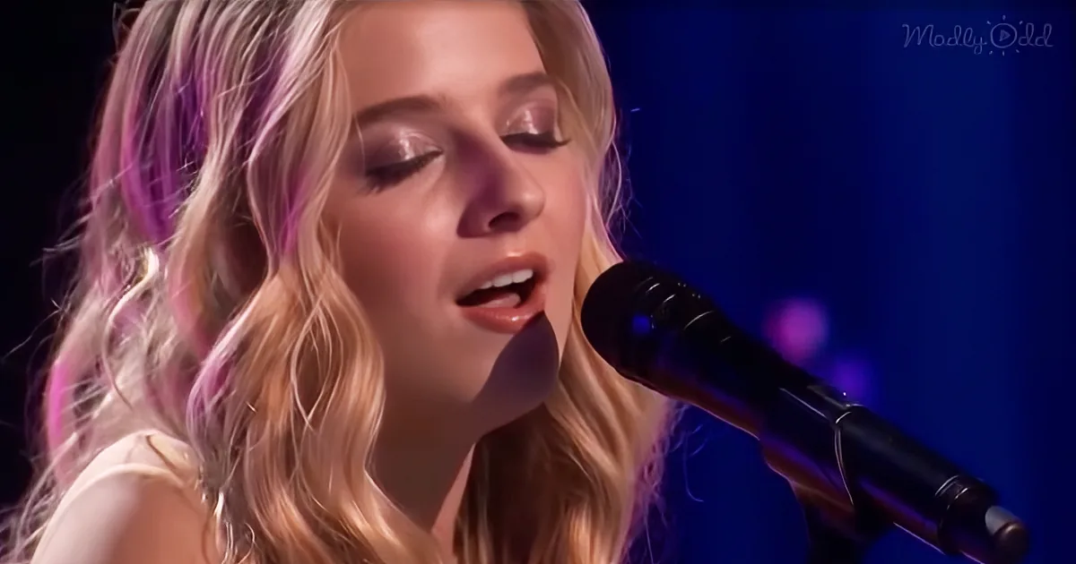 A woman with long blonde hair singing into a microphone, eyes closed, in a dimly lit setting.
