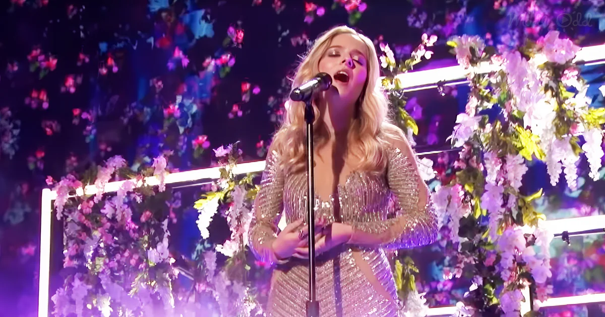 A woman in a sparkly dress sings into a microphone, surrounded by floral decorations.