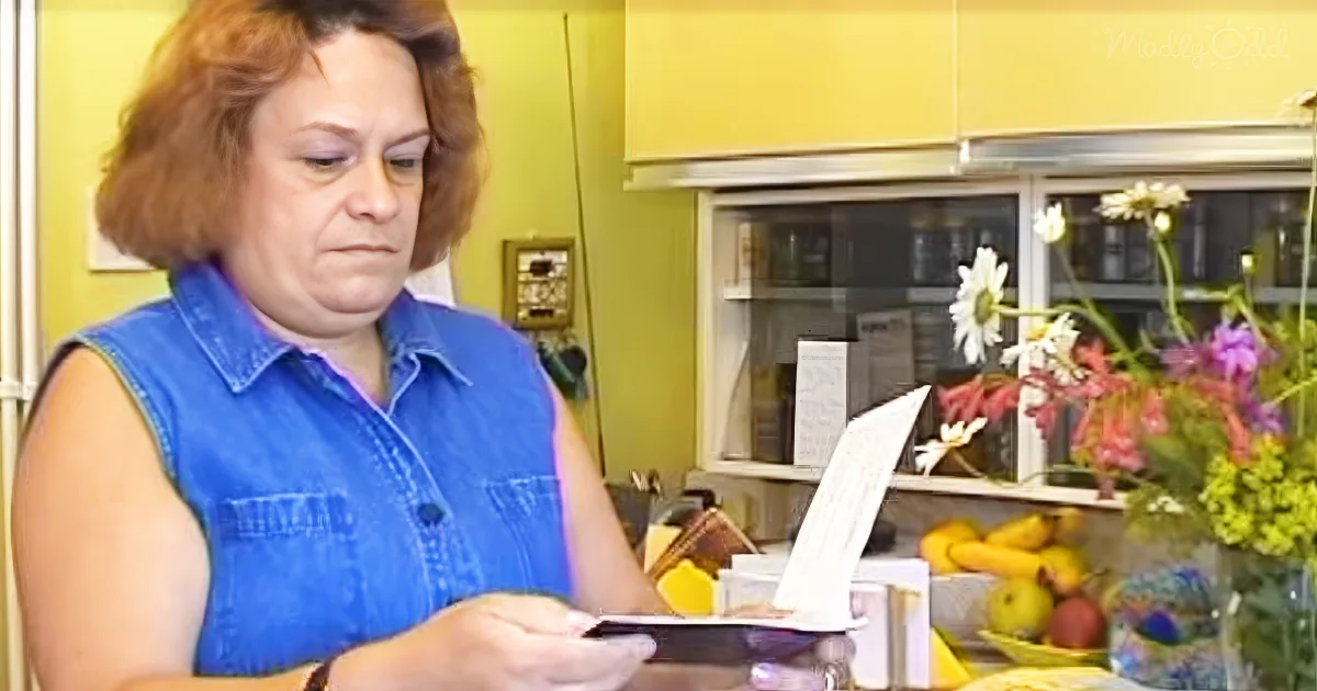 A woman in a blue shirt is holding a piece of paper, looking at it in a kitchen setting.