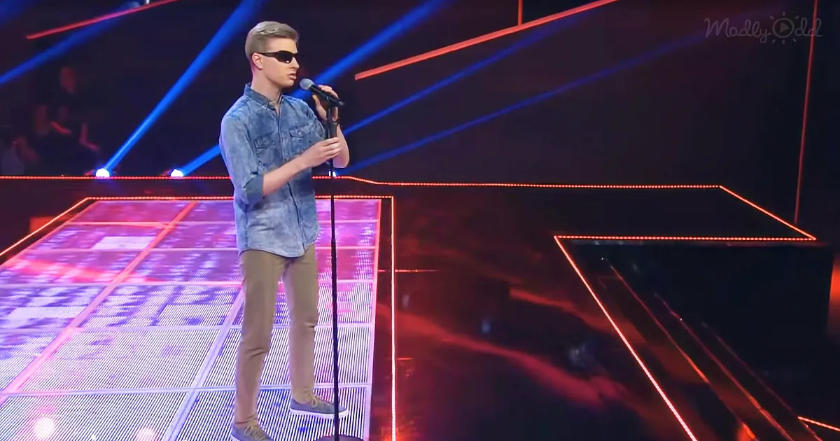 A man in sunglasses and a denim shirt stands on a stage with a microphone, illuminated by red and blue lights.