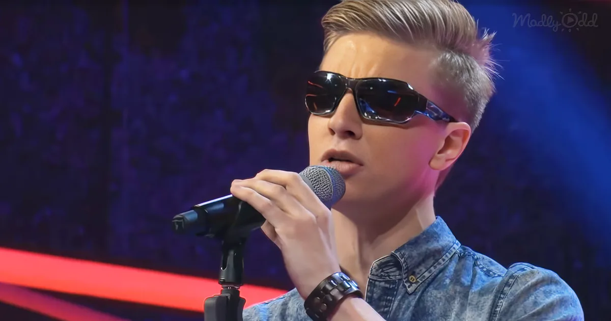 A person wearing sunglasses and a denim shirt sings into a microphone on stage.