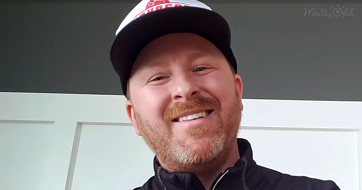A smiling man with a beard, wearing a black jacket and a white cap with red text, stands in front of a white door.