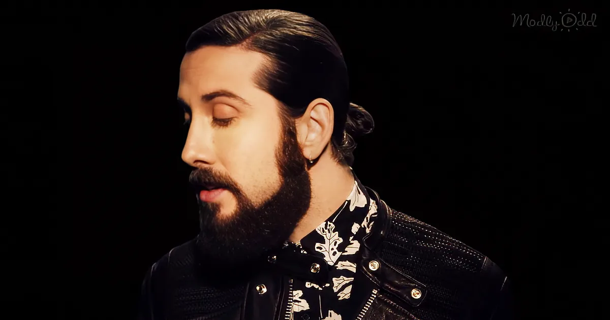 A man with a beard and ponytail, wearing a black leather jacket and a floral shirt, is seen in profile against a dark...
