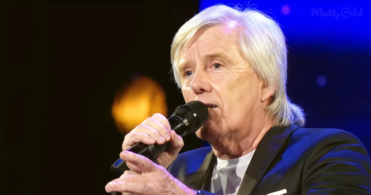An older man with white hair sings into a microphone, wearing a black suit and white shirt.