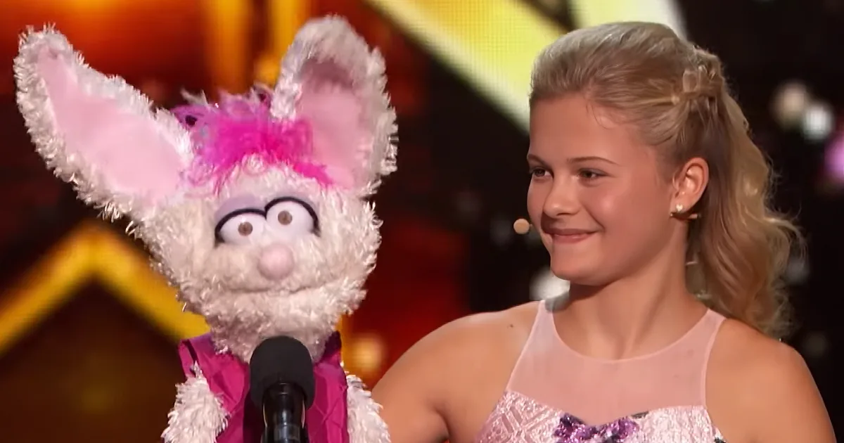 A young girl in a pink dress stands next to a white puppet with pink ears and a purple outfit, both on stage.