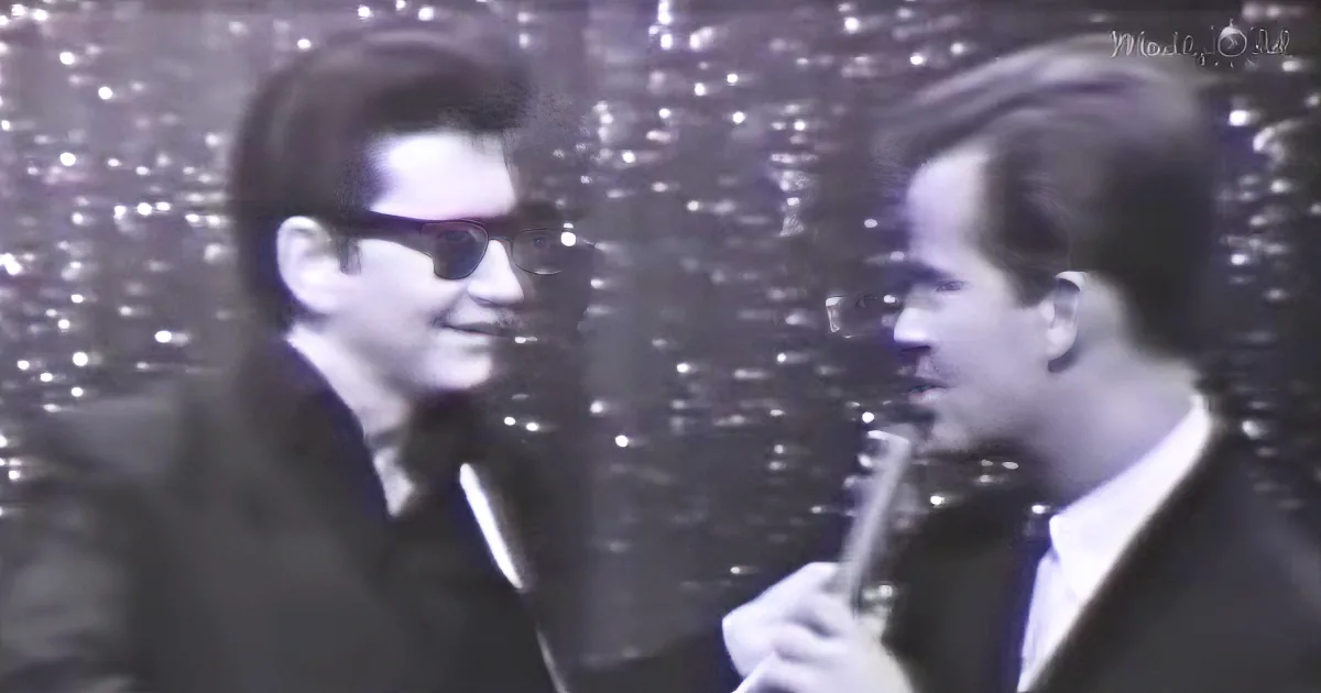 Two men in suits, one with sunglasses, holding microphones, standing in front of a glittery backdrop.