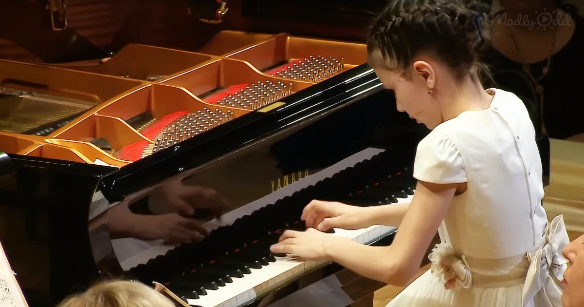A young girl in a white dress plays a Yamaha grand piano, with another person assisting.
