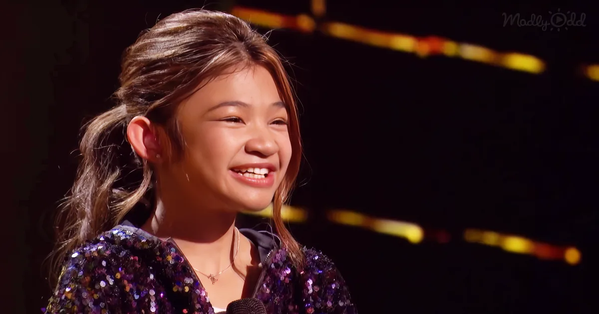 A smiling young girl with long hair, wearing a sparkly purple outfit, stands in front of a microphone on stage.