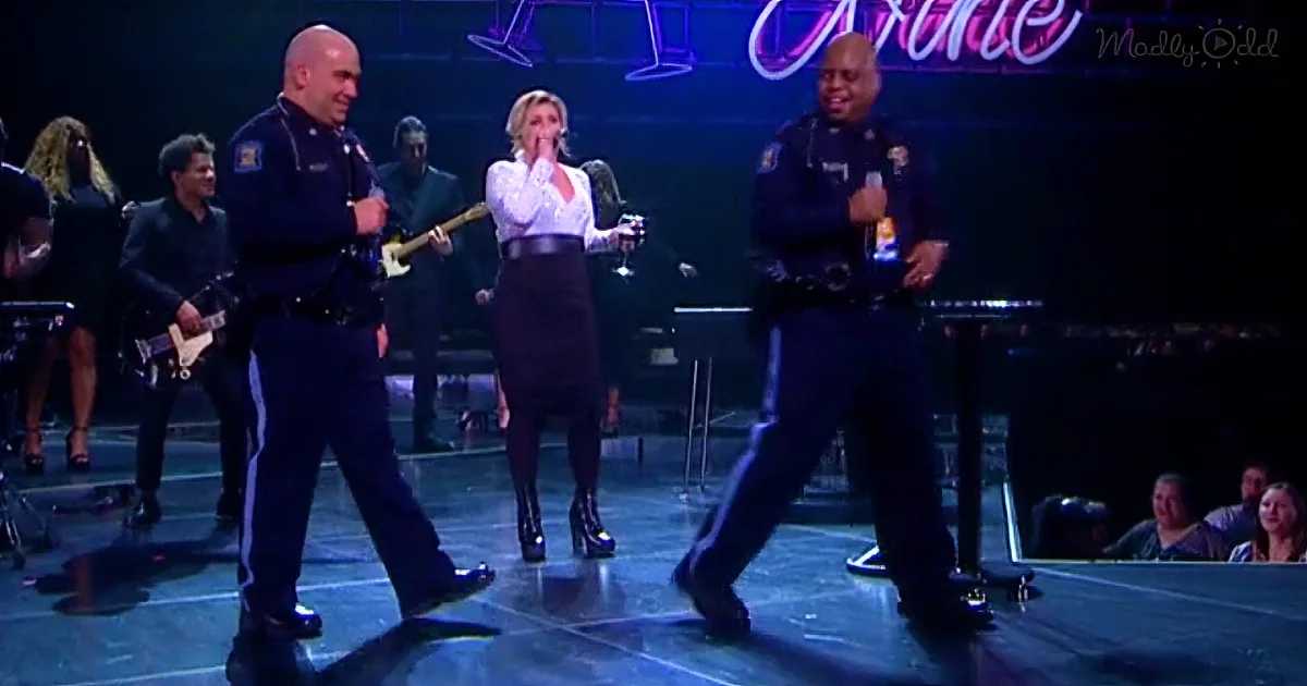 A woman in a white blouse and black skirt stands on stage with two police officers, surrounded by musicians.
