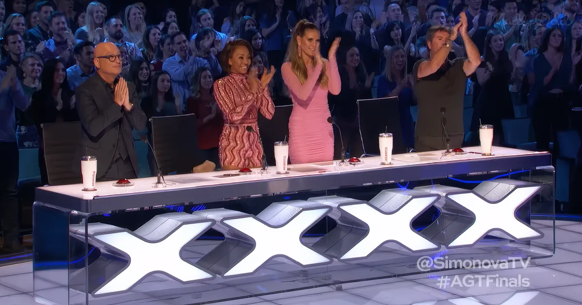 Judges and audience applauding at a talent show with "XXX" logo on the table.