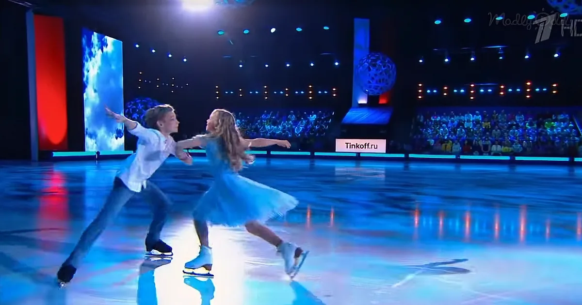 A couple in synchronized skating moves on ice, surrounded by a vibrant stage and audience.