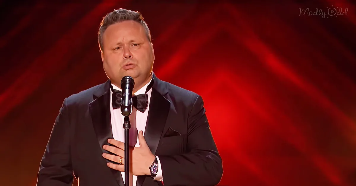 A man in a tuxedo sings into a microphone on stage with a red background.
