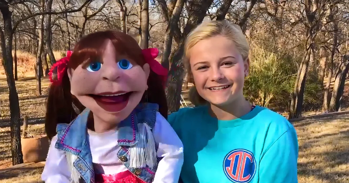 A girl in a blue shirt smiles next to a puppet with red hair and a denim jacket in a forest setting.