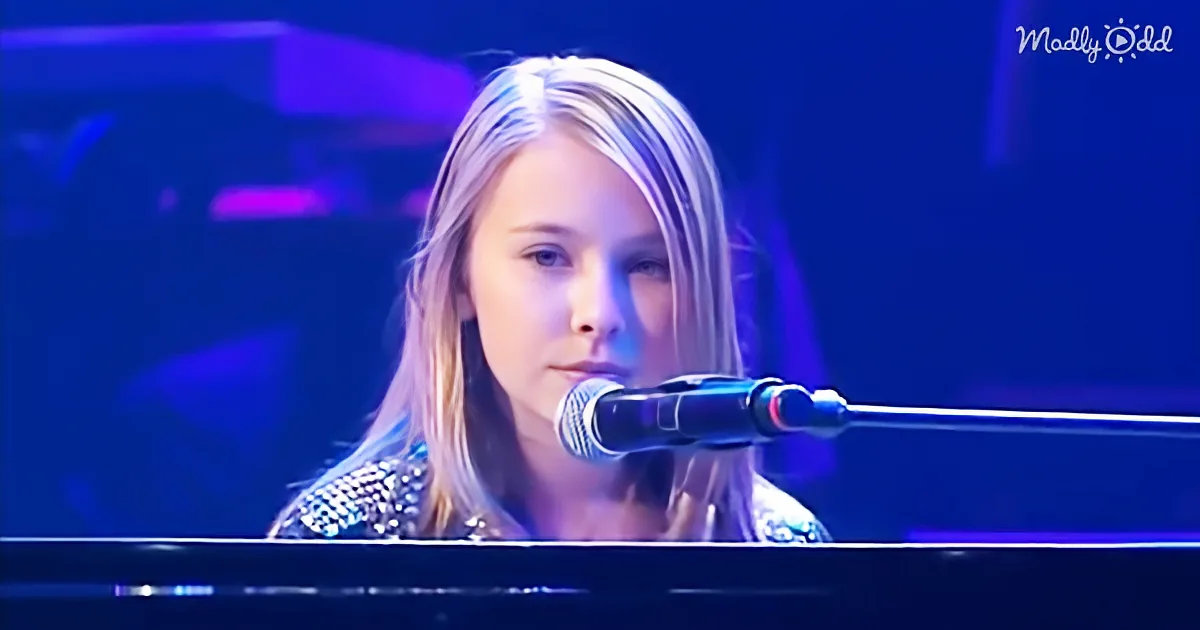 A young person with long hair sings into a microphone on stage under blue lighting.
