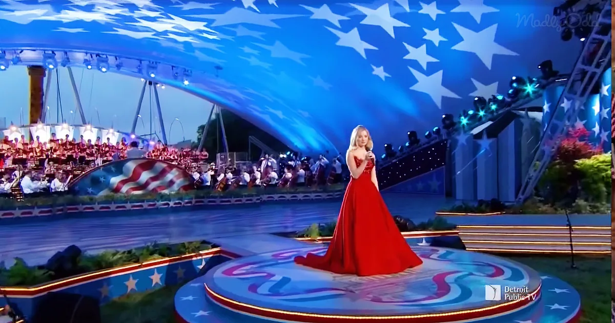 A woman in a red dress sings on a patriotic-themed stage with an orchestra and American flag backdrop.
