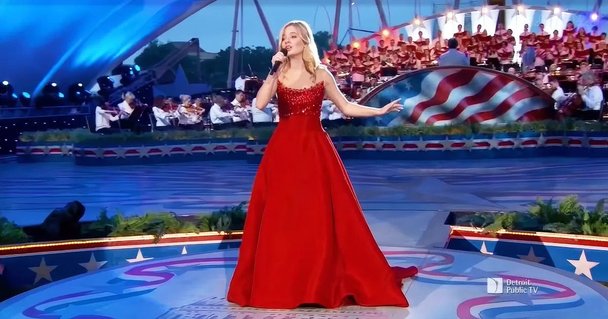 A woman in a red gown sings on stage with a large American flag backdrop and an orchestra.