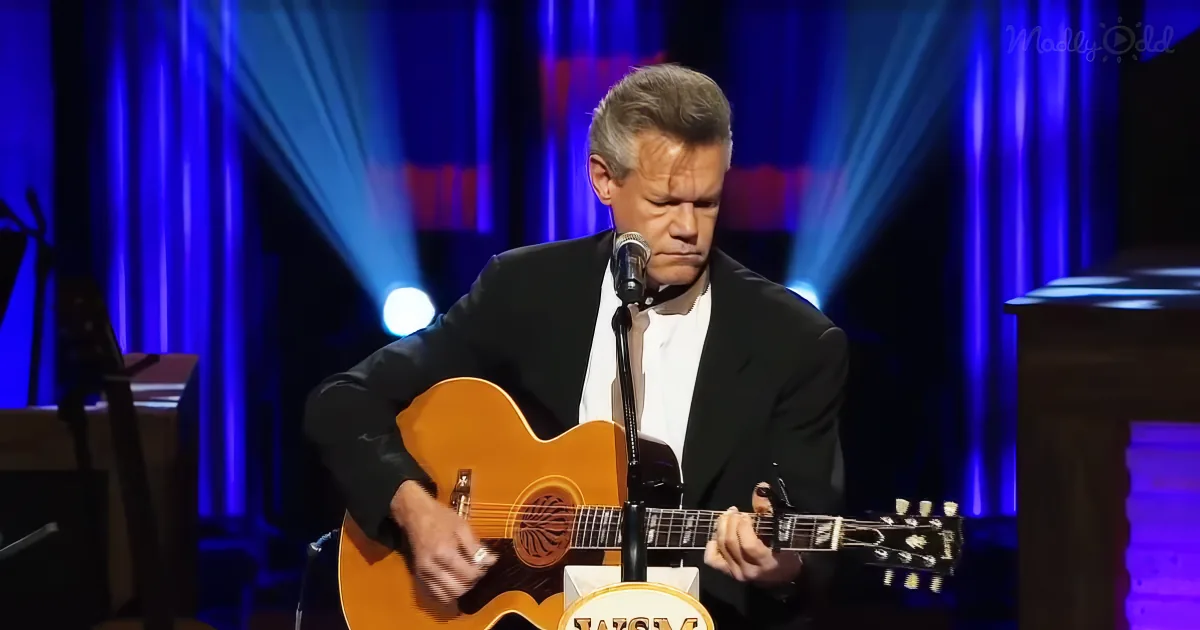 A man in a suit plays an acoustic guitar on stage with blue lighting.