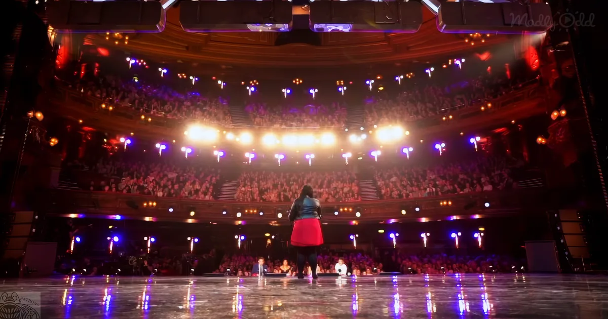 A person in a red skirt stands on a stage in front of a large, enthusiastic audience in a grand theater.