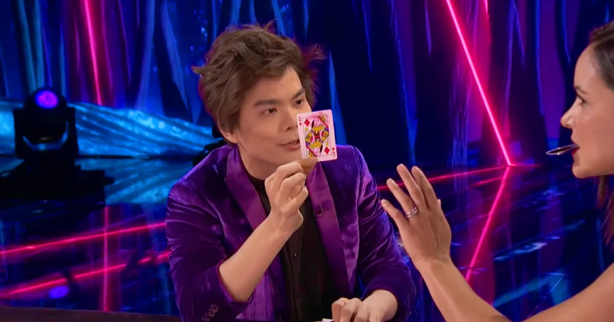 A man in a purple suit holds a card while a woman gestures with her hand in a colorful, stage-like setting.