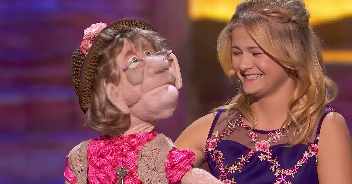 A puppet with glasses and a pink outfit is being held by a person in a floral dress, both smiling.