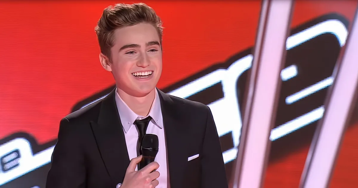 A young man in a black suit smiles while holding a microphone on stage.