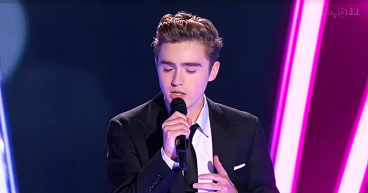 A young man in a black suit sings into a microphone on stage with vibrant pink and blue lighting.