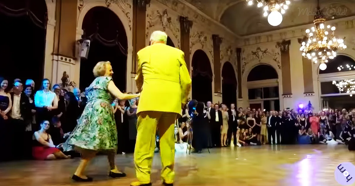 A man in a bright yellow suit dances with a woman in a floral dress in a grand hall with a chandelier.