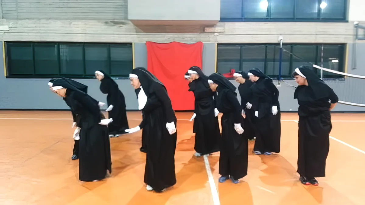 Nuns in black habits and white veils stand in a line on a gymnasium floor, heads bowed.