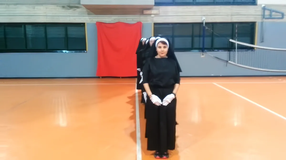 A person in a nun costume stands on an indoor court with a red cloth and a volleyball net in the background.