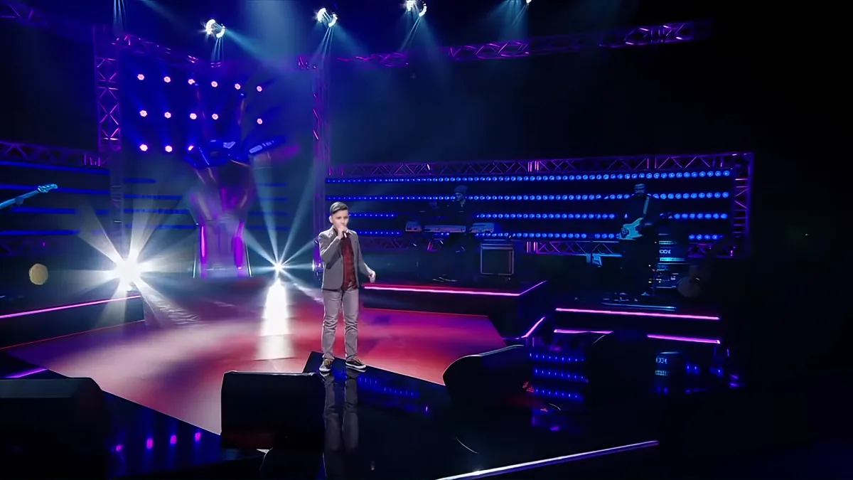 A person stands on a brightly lit stage with colorful lights and a large screen behind them.