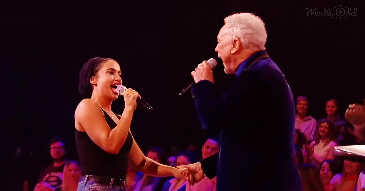 A woman and an older man sing together on stage, holding hands, with an audience watching.