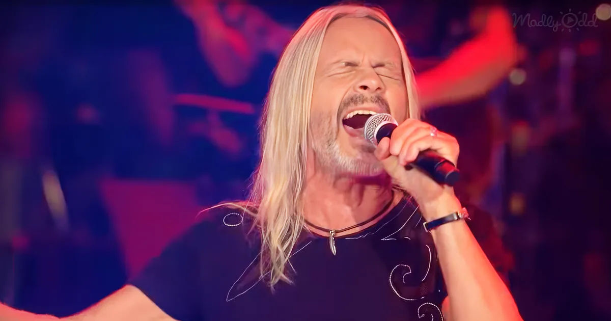 A man with long blonde hair sings passionately into a microphone on stage.