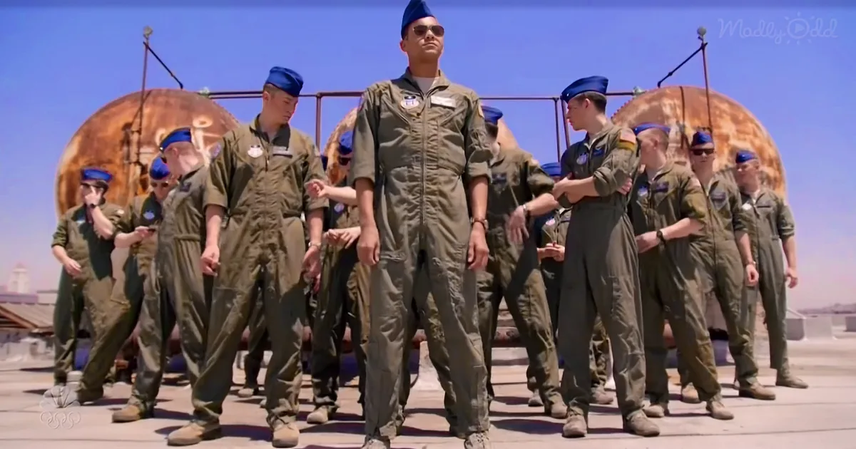 A man in a green flight suit stands in the center, surrounded by others in similar attire, on a rooftop under a clear sky.