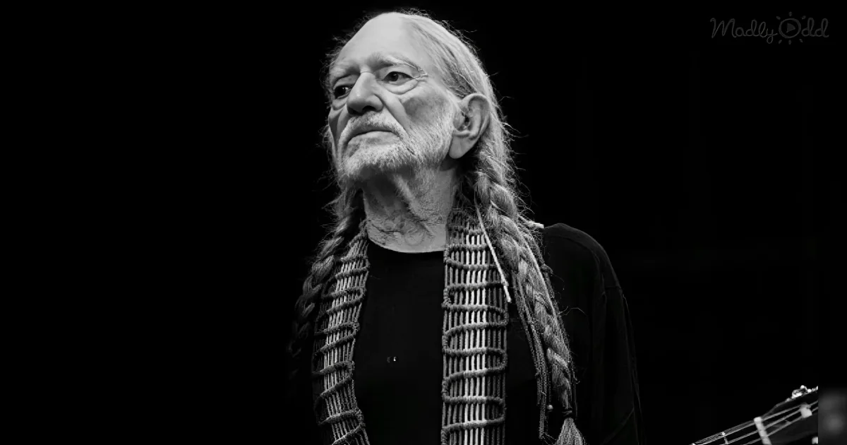 A man with long braided hair and a beard, wearing a black shirt, holding a guitar, against a dark background.