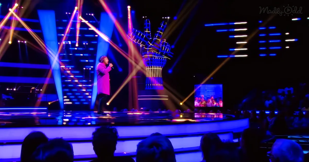 A person in a pink coat sings on a stage with vibrant blue and yellow lights.