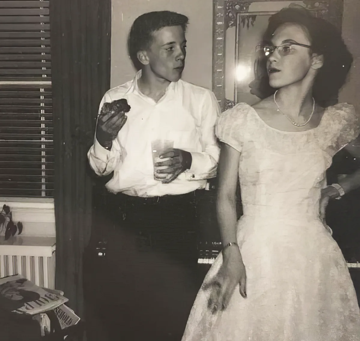 A man in a white shirt and dark pants holds a drink, while a woman in a white dress and glasses stands beside him, both...