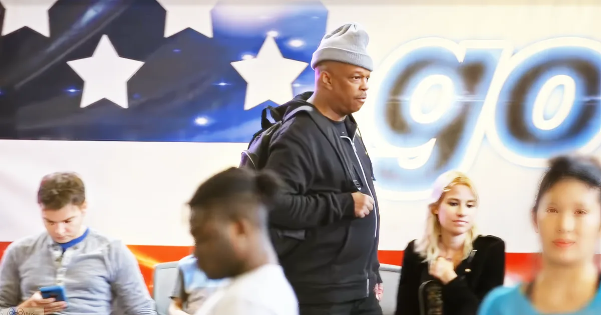 A man in a beanie and black jacket walks past a large American flag and "GO" sign.