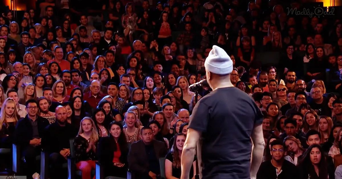 A man in a white beanie and black shirt stands on stage, facing a large audience of smiling and engaged spectators.