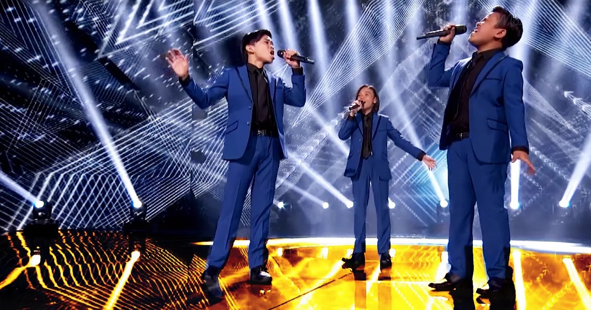 Three performers in blue suits sing on stage with dynamic lighting and a futuristic backdrop.