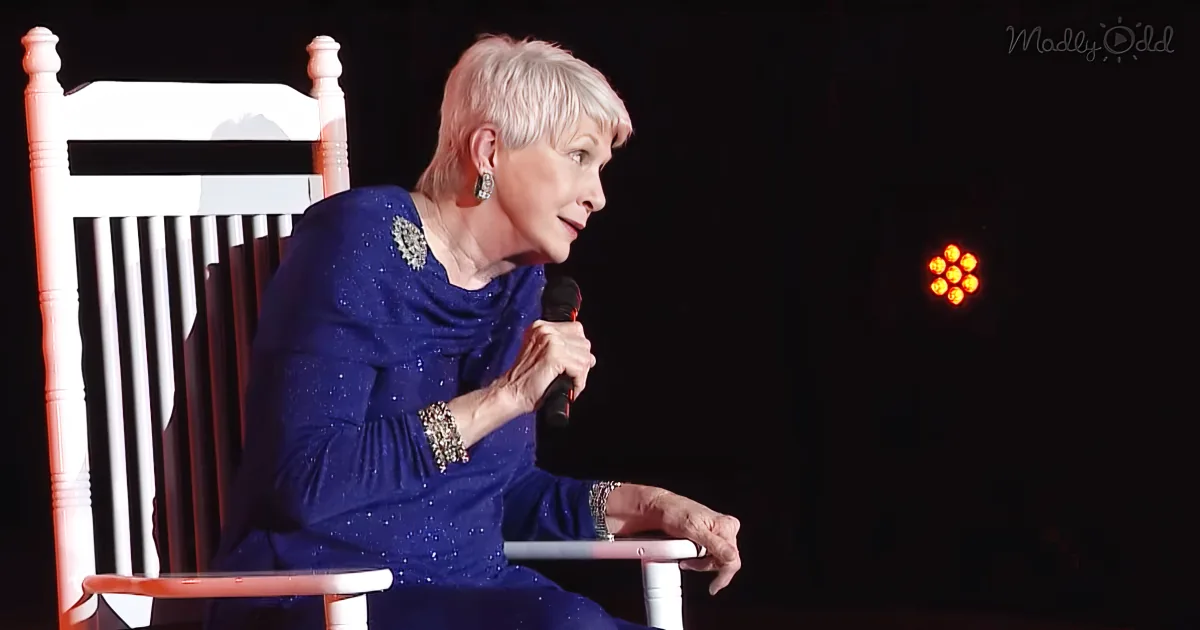 An older woman in a blue dress sits on a white rocking chair, holding a microphone, with a red light in the background.